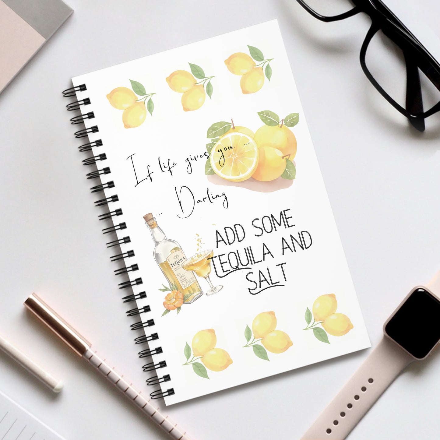 Notebook with lemon design and humorous quote on a white surface with glasses and a smart watch and some stationery.